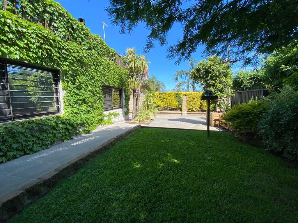 A serene outdoor space is framed by lush greenery and splashes of sunlight. A well-maintained lawn extends across the area, with garden features such as palm trees and shrubs visible. Pathways are lined with natural stone, leading to a secluded seating area.