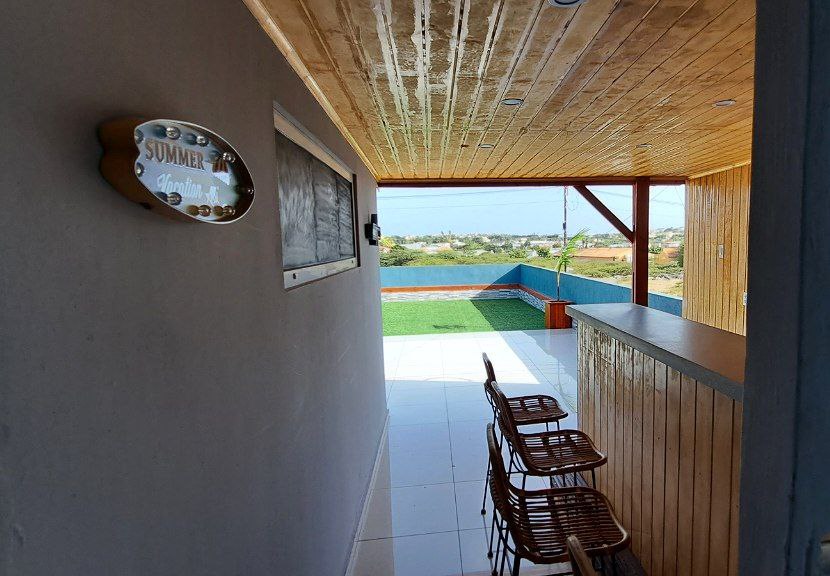 An outdoor terrace is visible, featuring a bar-like area with several wooden stools. A scenic view of the landscape and water can be seen through the open space, with well-maintained green grass observed in the distance. A sign indicating 'SUMMER' is mounted on the wall.