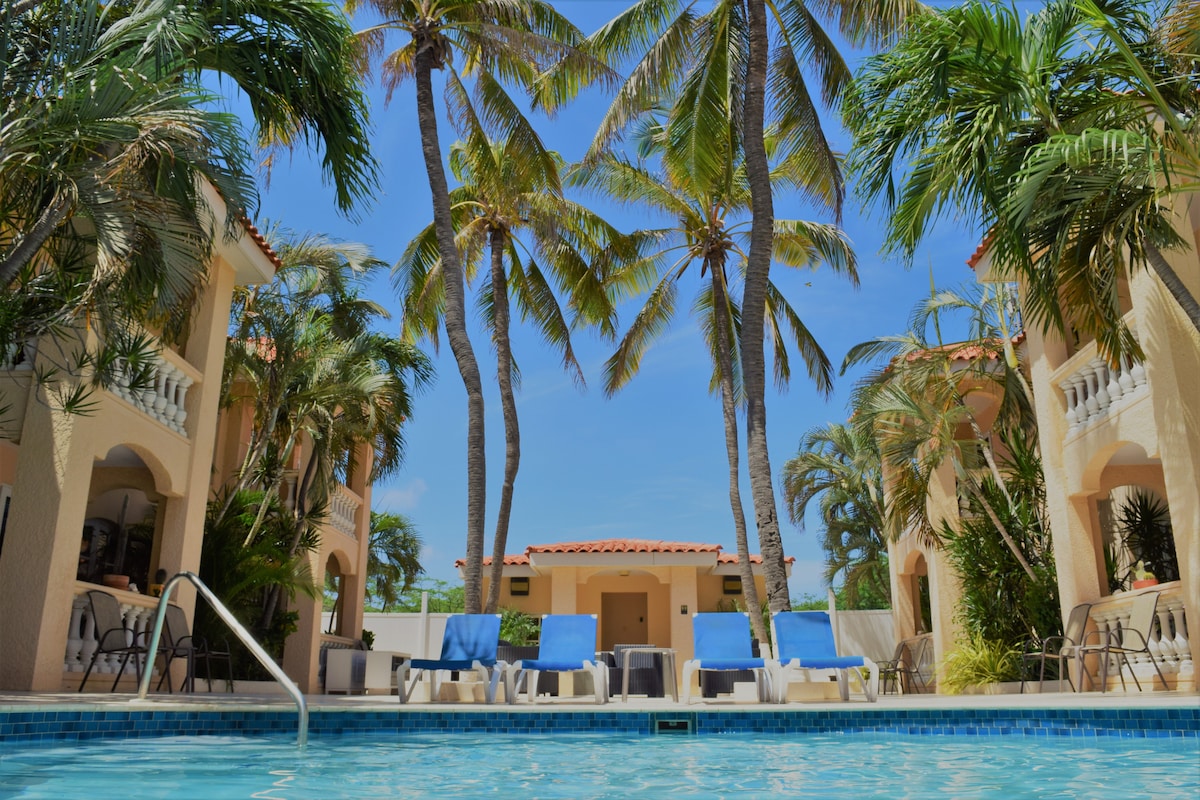 The image captures a tranquil pool area surrounded by tall palm trees and lush greenery. Sun loungers are arranged by the pool, with the condominium's architecture visible in the background, showcasing a blend of vibrant colors and outdoor spaces.