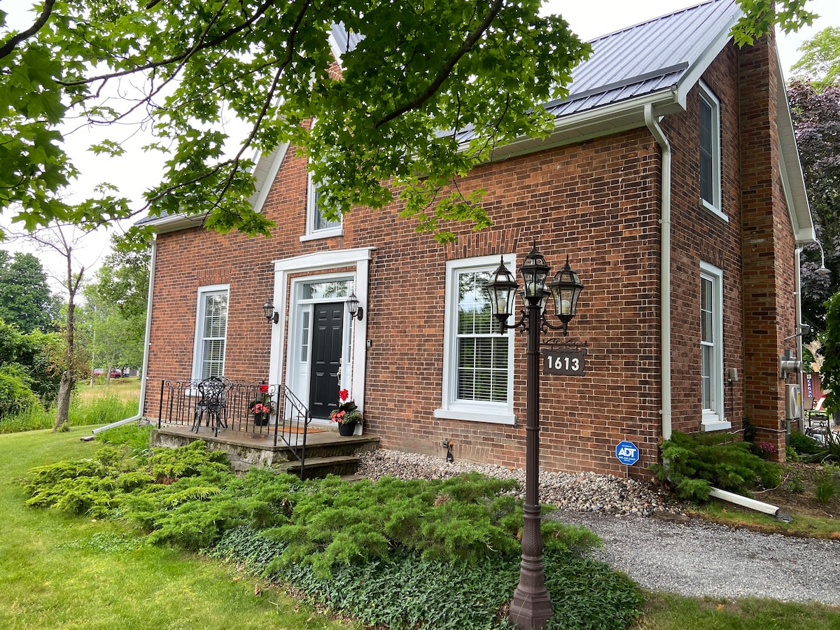 The exterior of the farmhouse features a classic brick façade with white trim. A welcoming front porch is adorned with potted flowers, while a pathway leads through well-maintained greenery. A charming street lamp stands nearby, adding to the inviting atmosphere.