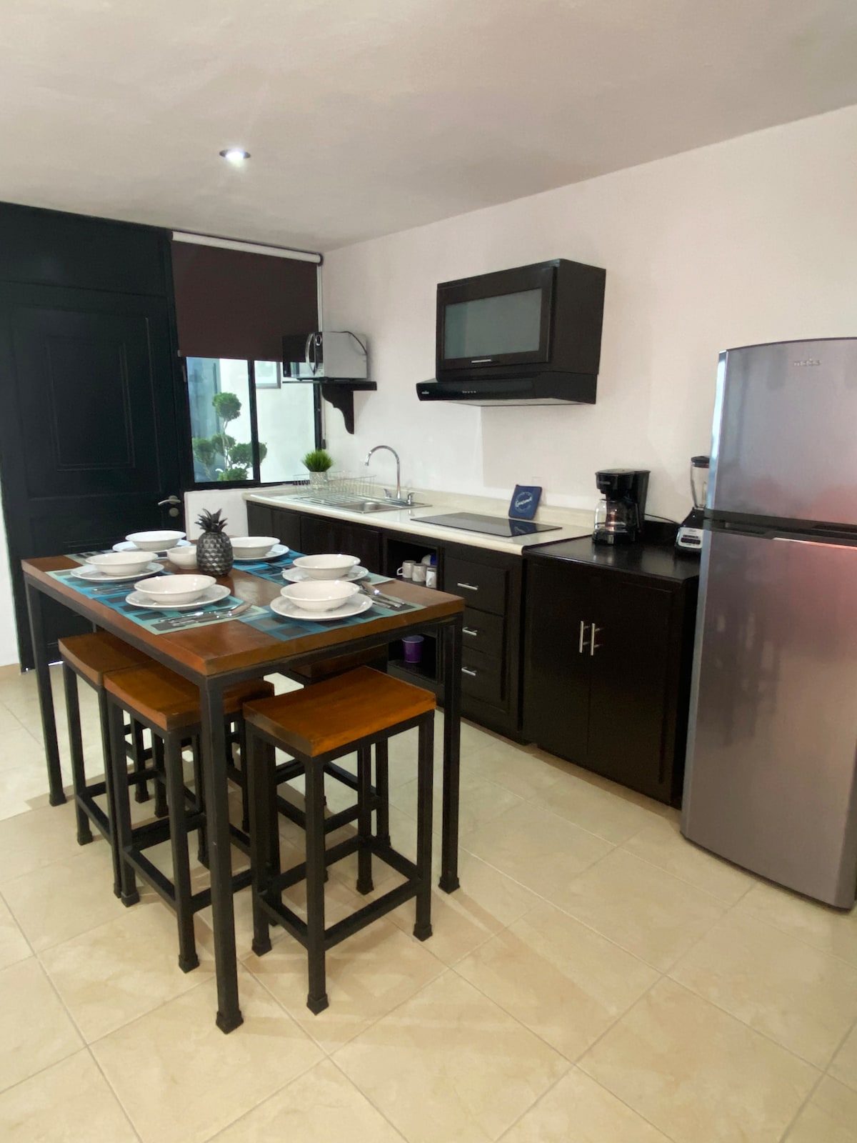 The kitchen area is equipped with modern appliances, including a refrigerator and microwave. A dining table with four wooden stools is set with dinnerware. Counter space is available, complemented by a sink and coffee maker, ensuring convenience for meal preparation.