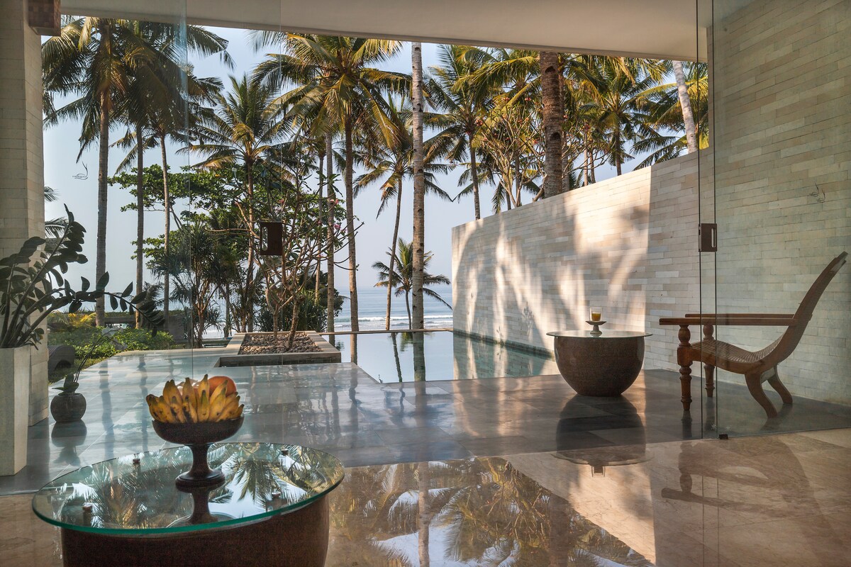 The image showcases a sleek interior that opens to a tranquil outdoor space, featuring a private pool surrounded by tall palm trees. A decorative bowl rests on a glass table, while a wooden chair complements the serene atmosphere.
