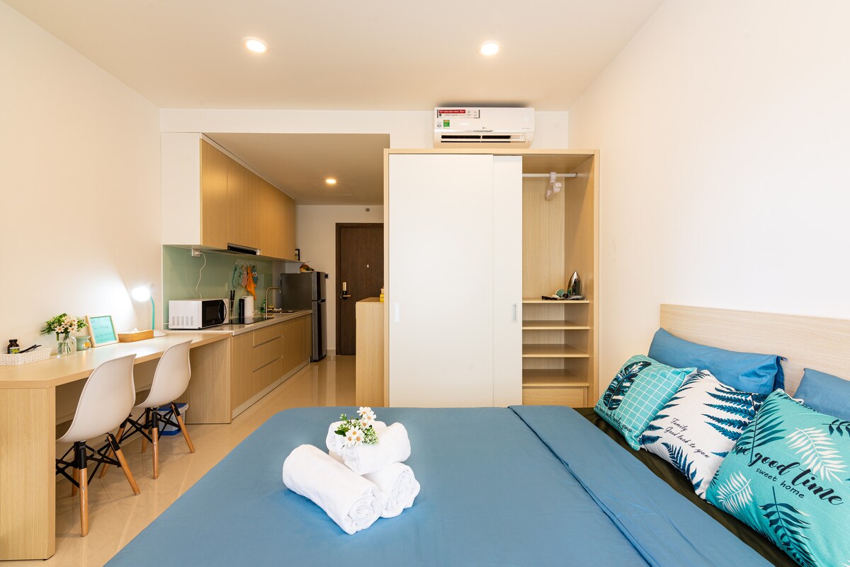 A spacious studio featuring a large, neatly made bed with decorative pillows. A kitchenette with cooking appliances is visible, along with a desk and chairs perfect for working. Fresh towels are neatly arranged on the bed, creating a clean and inviting environment.