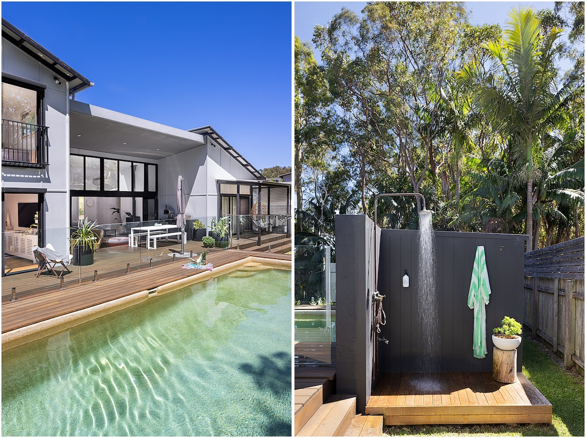 A modern beach house features a spacious deck overlooking a clear swimming pool, flanked by lush greenery. Large glass doors lead to an open living space, inviting natural light. An outdoor shower stands nearby, surrounded by privacy screening and tropical plants, with a towel hanging ready for use.
