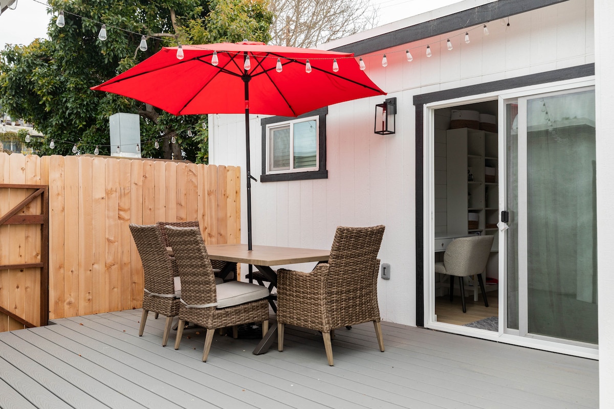 A spacious outdoor deck features a large red umbrella providing shade over a dining table surrounded by four wicker chairs. Soft lighting strings are suspended overhead, adding warmth to the area. A sliding glass door leads to an adjoining indoor space.