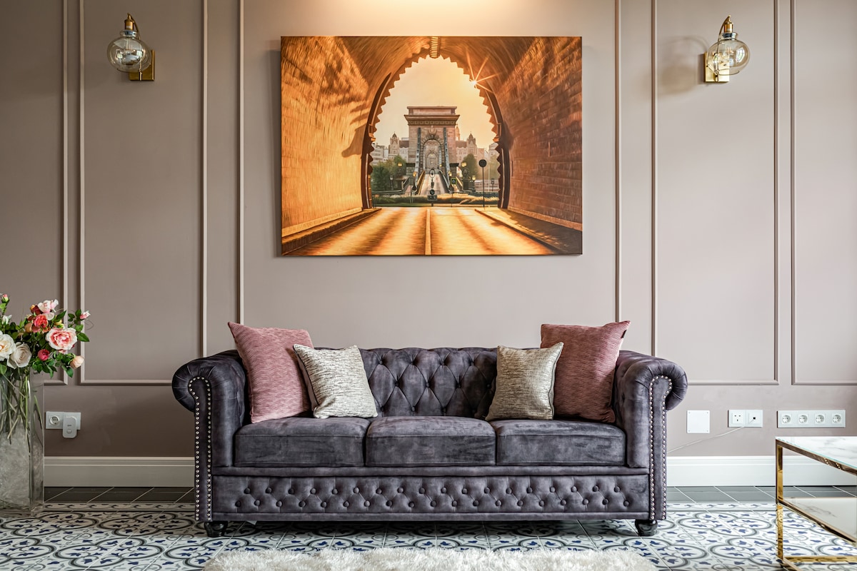 A Chesterfield-style couch in a rich shade of purple is positioned against a softly lit wall. Decorative pillows in subtle pink hues add comfort. Above, a large artwork depicts an inviting archway, while a modern light fixture illuminates the space, enhancing the elegant atmosphere.