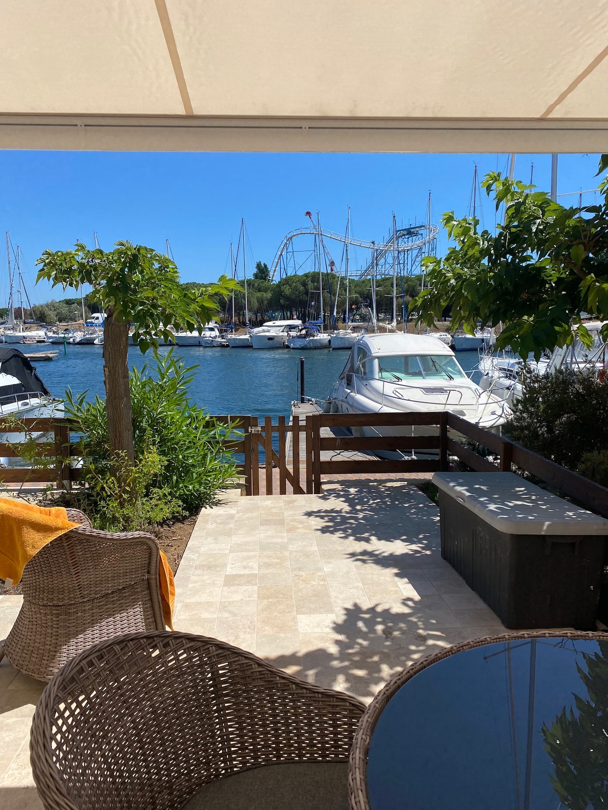 A comfortable patio area features two woven chairs and a small table, providing a view of the marina and docked boats. Sunlight filters through the retractable awning, creating a pleasant space for relaxation. Lush greenery surrounds the patio, adding to the serene ambiance.