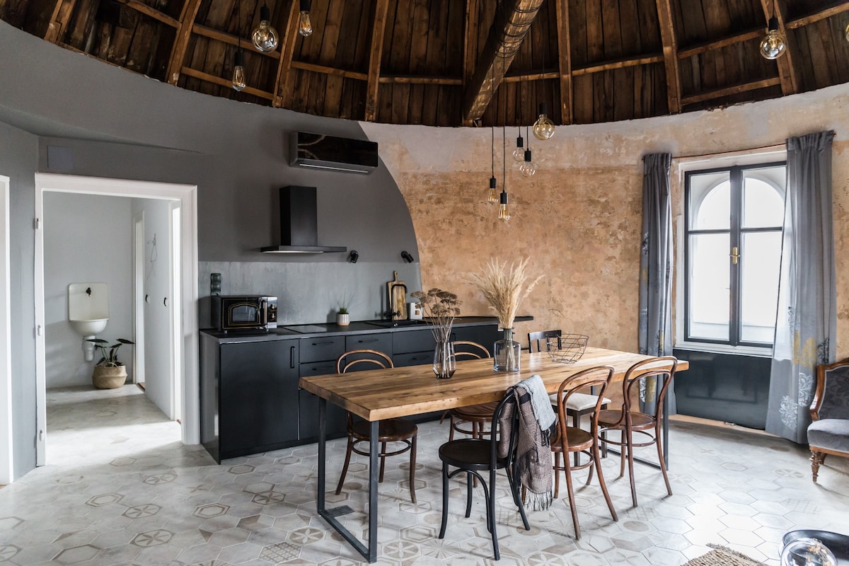 An open dining area features a rustic wooden table surrounded by several chairs, complemented by a dark cabinetry and modern appliances. Soft lighting hangs from the circular ceiling, and large windows with sheer curtains allow natural light to fill the space.