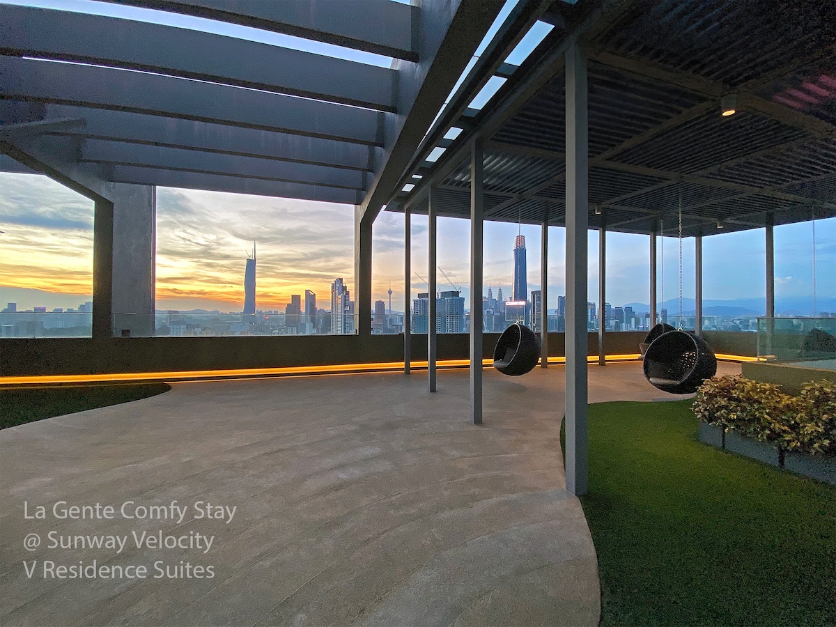 An open rooftop space offers a panoramic view of the Kuala Lumpur skyline, featuring modern buildings and a sunset sky. Two hanging chairs provide a relaxing spot to enjoy the surroundings, surrounded by greenery and ambient lighting along the pathway.