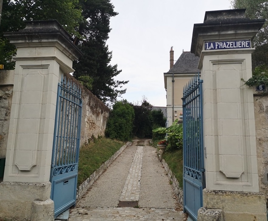 The image features an entrance gate marked 'La Frazeliere,' flanked by tall stone pillars. A gravel pathway leads into the property, bordered by greenery. The path is lined with stones, guiding visitors toward the house that is partially visible to the right.