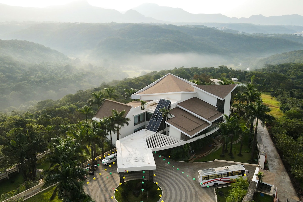An aerial view captures the villa nestled among lush greenery, showcasing its modern architecture and expansive layout. Solar panels adorn the roof, and the circular driveway is lined with palm trees, providing a sense of tranquility amidst the scenic landscape.
