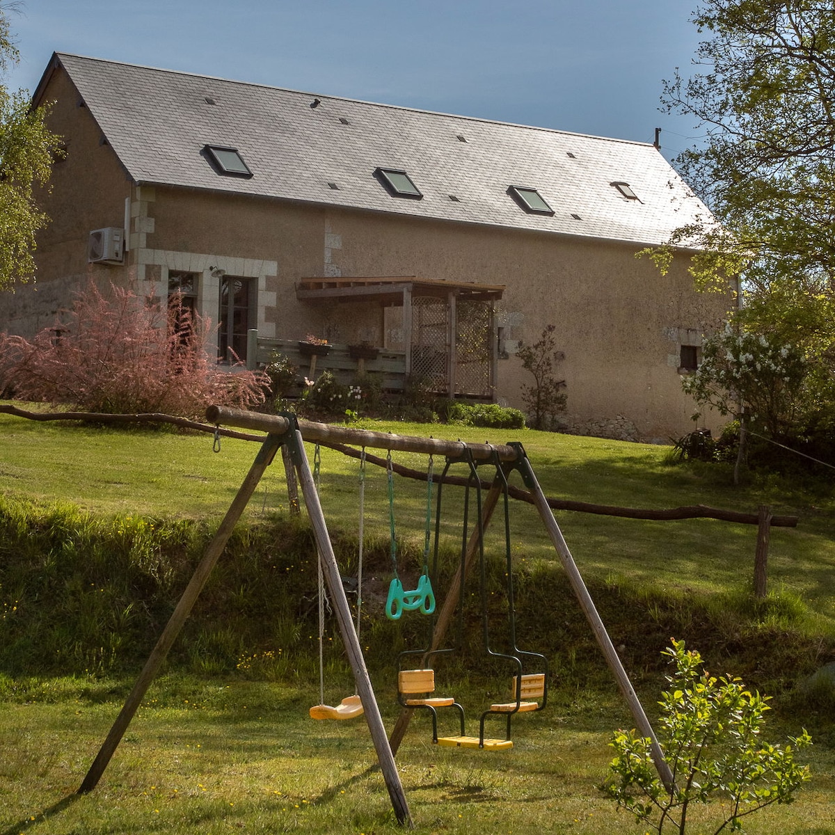 A spacious garden area features a wooden swing set with two swings, surrounded by lush green grass. The house is visible in the background, showcasing a simple, rustic design under a clear blue sky.