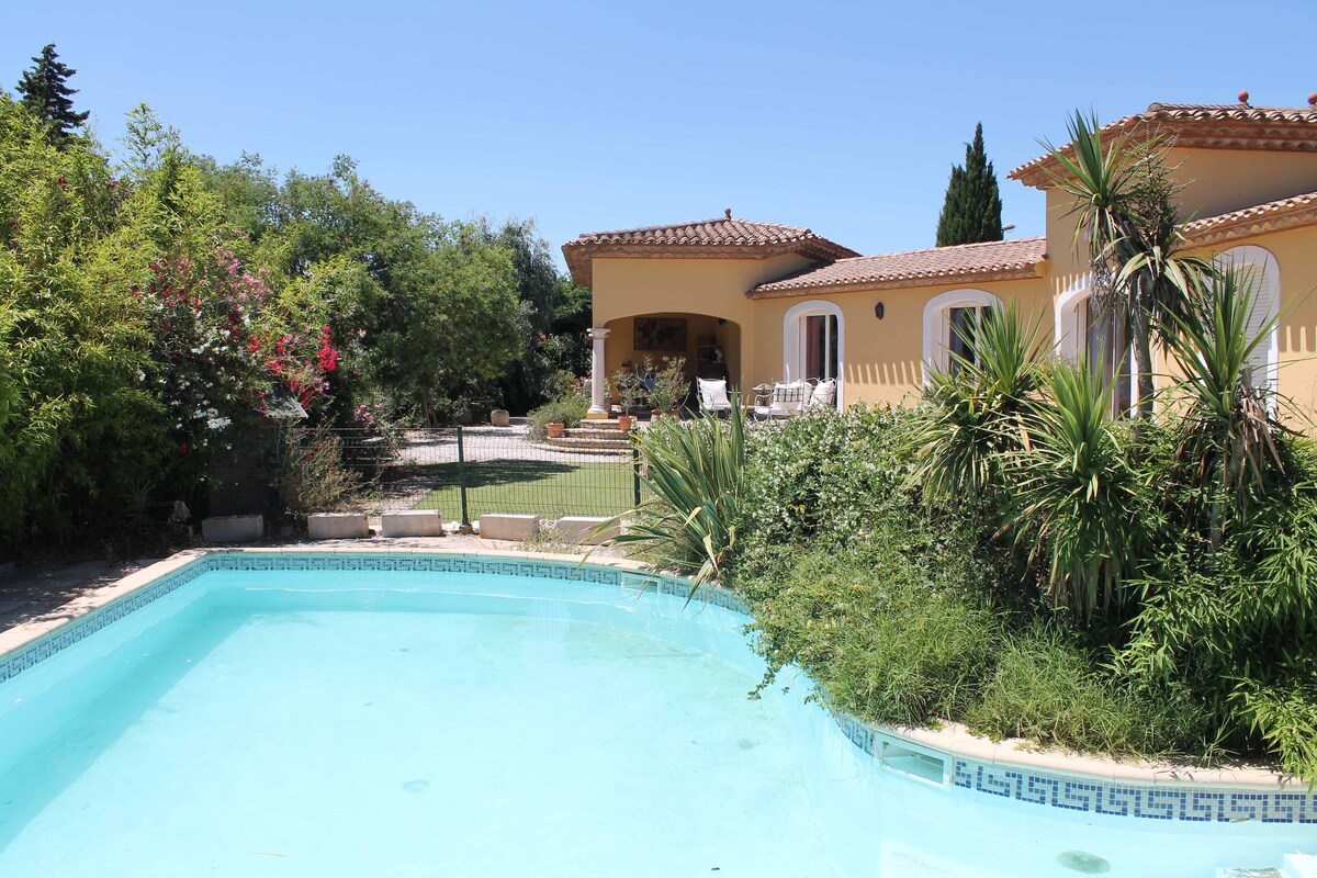 A clear swimming pool is surrounded by lush greenery and flowering plants. The villa's exterior is visible with a tiled roof and multiple windows. A terrace area is seen adjacent to the pool, providing a serene space for relaxation.