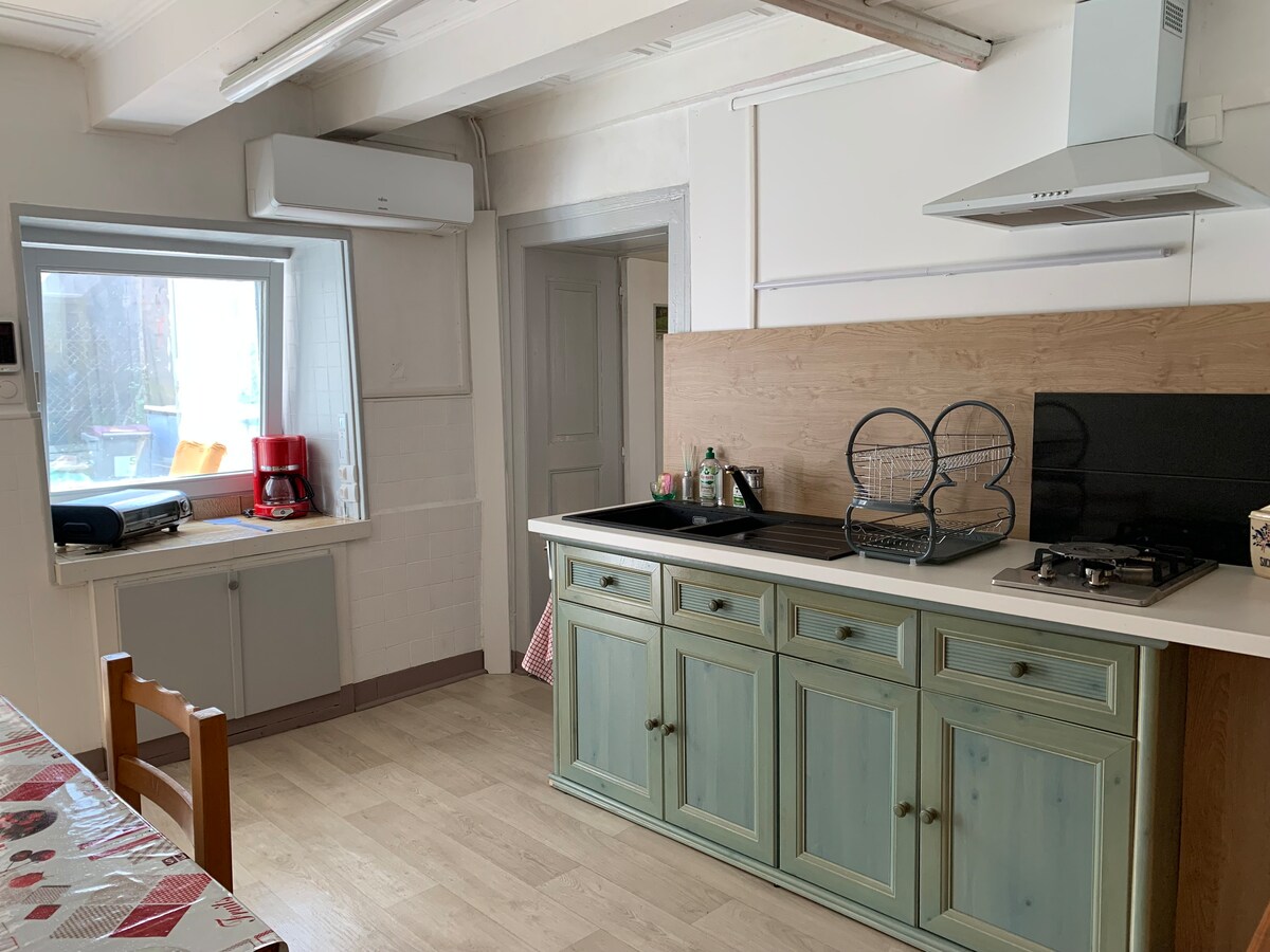 A bright kitchen area is featured, showcasing light-colored cabinetry and a stovetop. A large window allows natural light to fill the space, complementing the wooden accents. Basic kitchen appliances are neatly arranged, contributing to a functional environment.