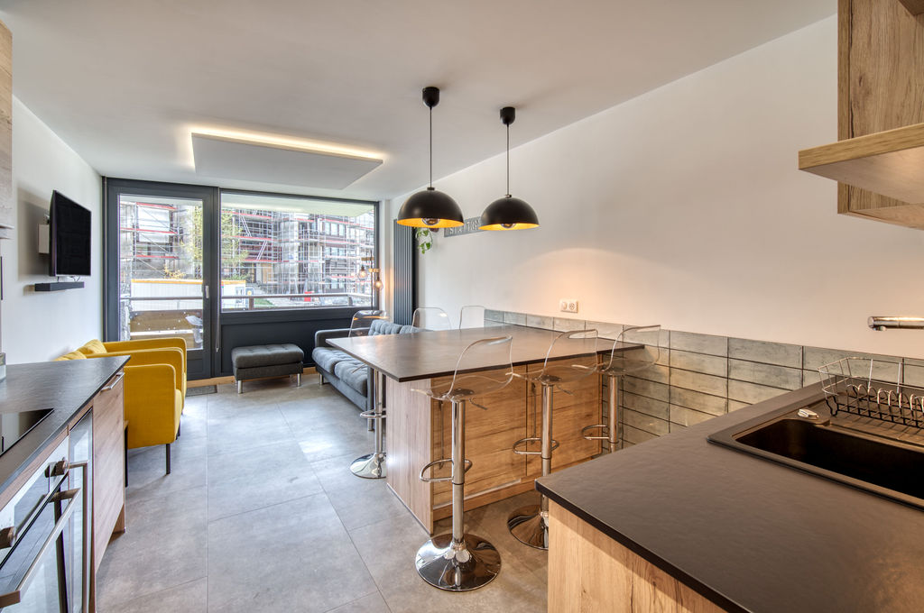A modern kitchen area features sleek cabinetry and stainless steel appliances. A spacious countertop is complemented by high stools. Large windows provide natural light and views of the outdoors. A cozy seating area includes a gray sofa and a yellow accent chair, creating a functional and inviting space.