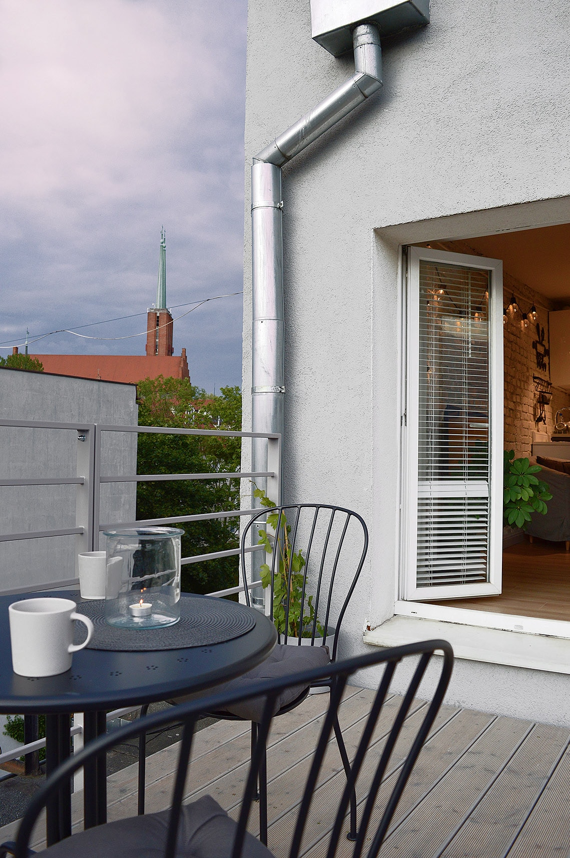 A balcony space features a small round table and two black chairs, accompanied by a clear glass jar. The room is accessible through a white-framed window, while a verdant tree provides a natural view. A church steeple is visible in the background under a cloudy sky.