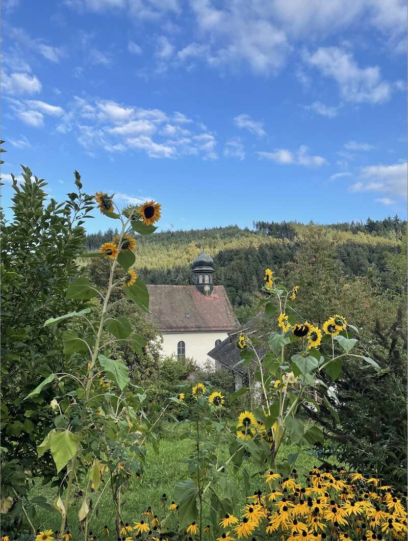 A village church is seen in the background, framed by vibrant sunflowers and greenery. The church features a distinctive steeple, and the sky is partially covered by clouds, creating a serene atmosphere. The surrounding landscape includes rolling hills and trees.