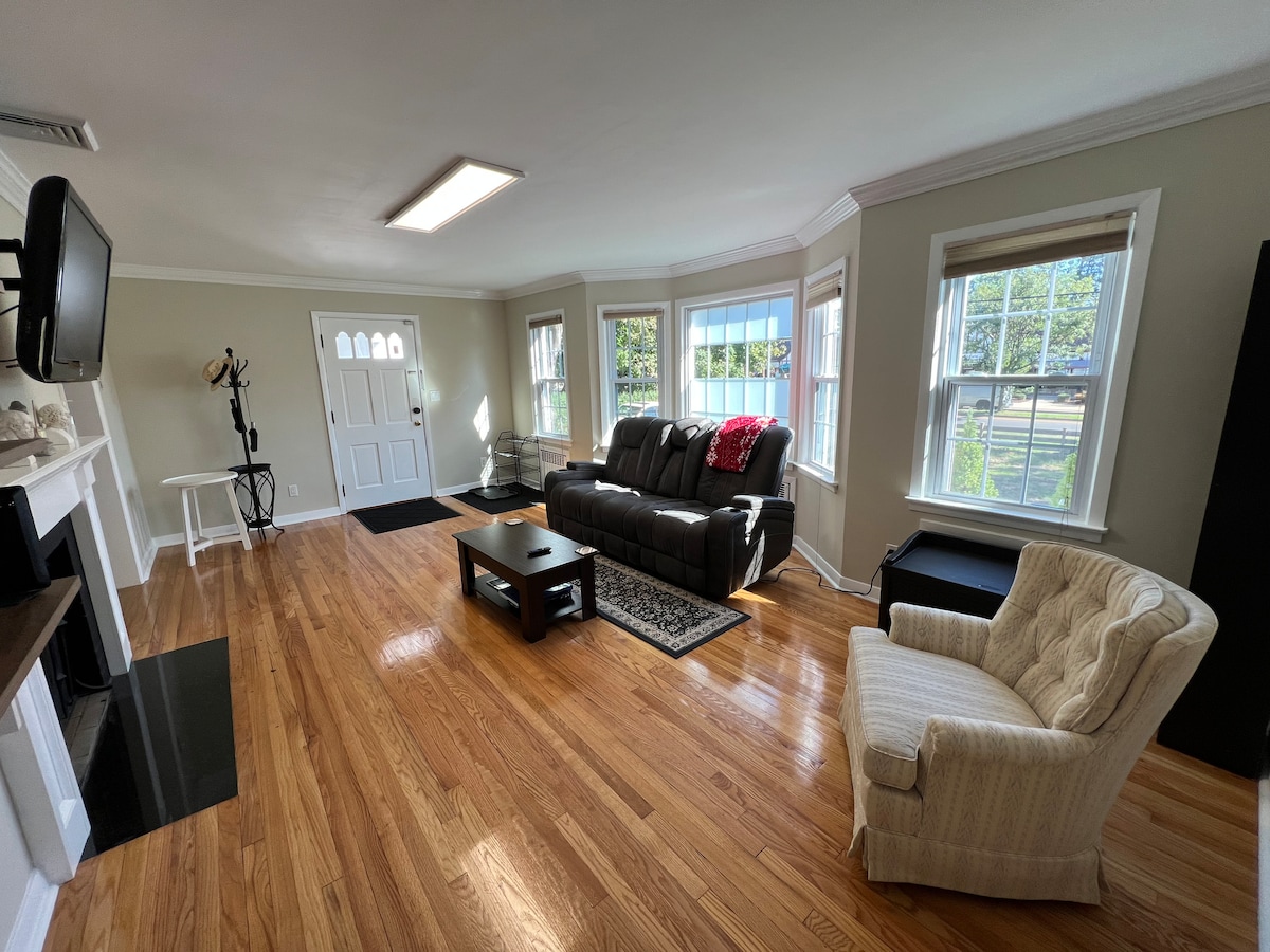 The living room features polished hardwood floors and an arrangement of comfortable seating. A recliner sofa is positioned alongside a classic armchair. Natural light floods in through multiple windows, illuminating the space. A flat-screen TV is mounted above a fireplace, with a coffee table situated in front.