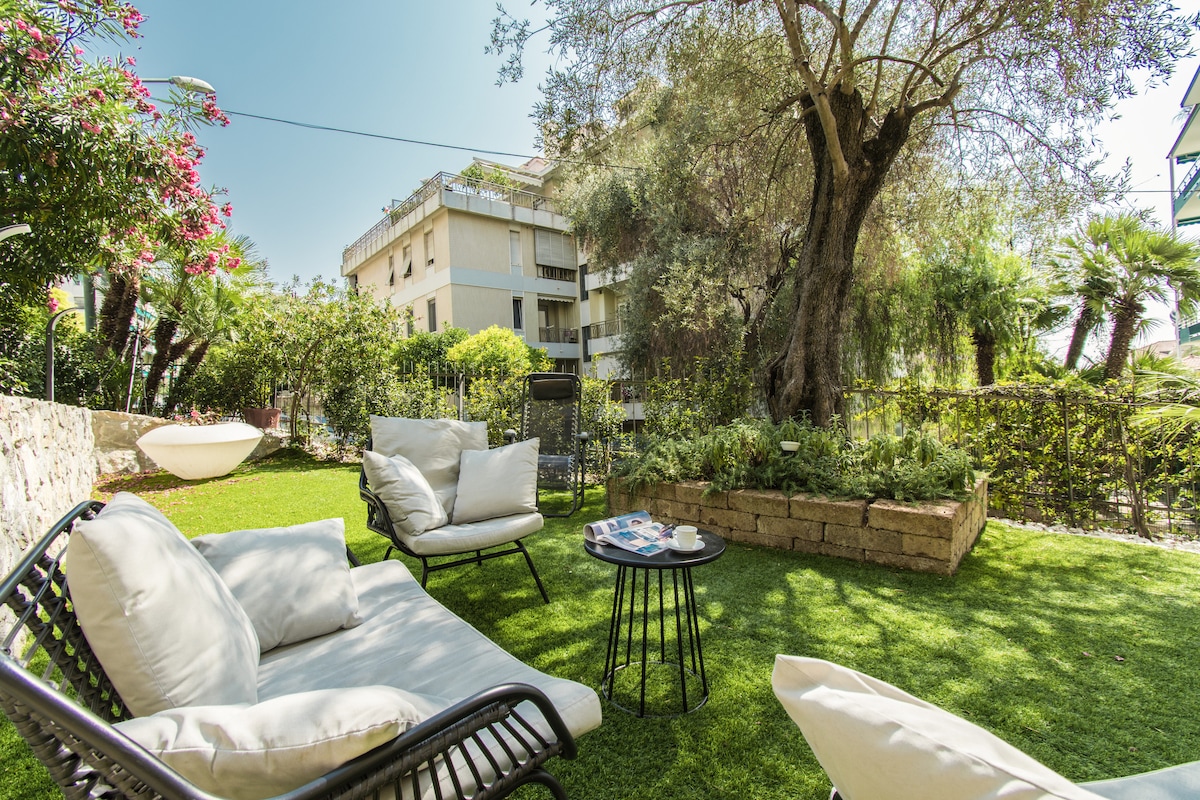 A garden area features comfortable lounge chairs positioned on vibrant green grass, accompanied by a small round table. Lush greenery, including trees and flowering plants, surrounds the space, providing a serene outdoor environment adjacent to the elegant condominium.
