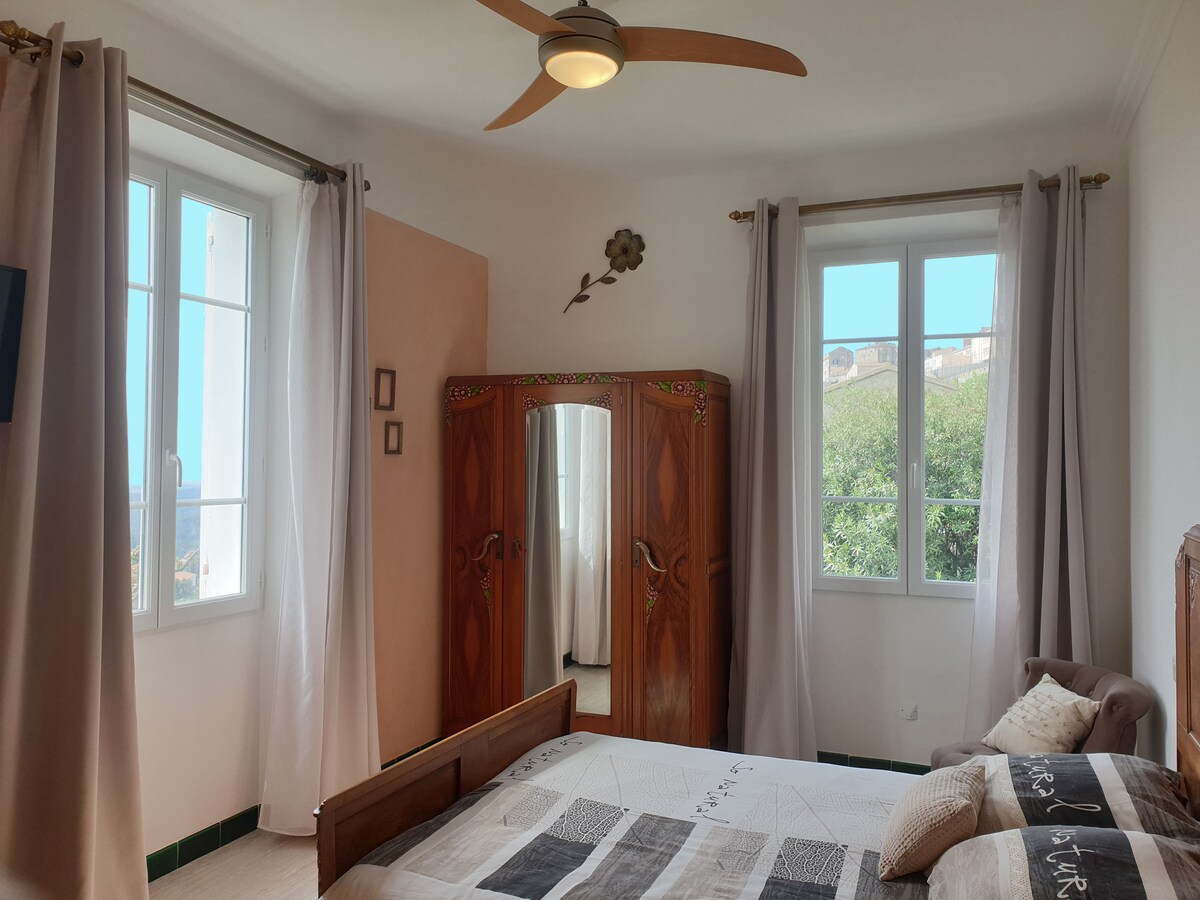 A warmly decorated guest room features a spacious bed with a textured bedding design. A wooden wardrobe with intricate detailing is positioned adjacent to the bed. Natural light enters through two large windows adorned with light curtains, highlighting the serene atmosphere.