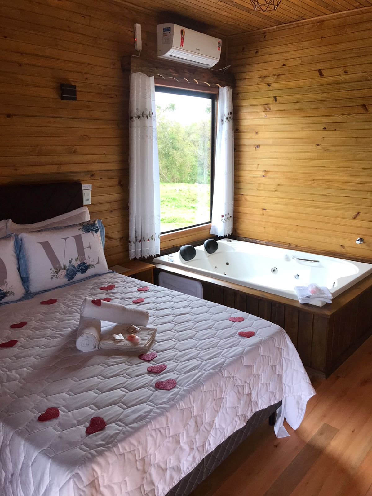 A wooden interior features a spacious bed adorned with decorative accents and a romantic arrangement of heart-shaped items. Beside the bed, a heated jacuzzi provides an inviting place to relax, while ample natural light enters through the window adorned with sheer curtains.