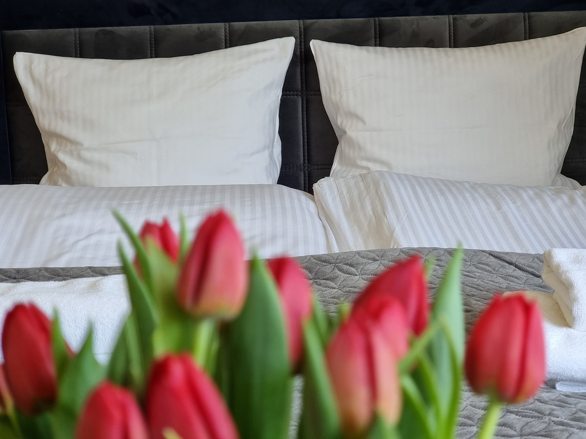 A spacious bed is positioned against a dark wall, featuring two neatly arranged white pillows with striped covers. In the foreground, a bouquet of vibrant red tulips adds a touch of color to the scene.