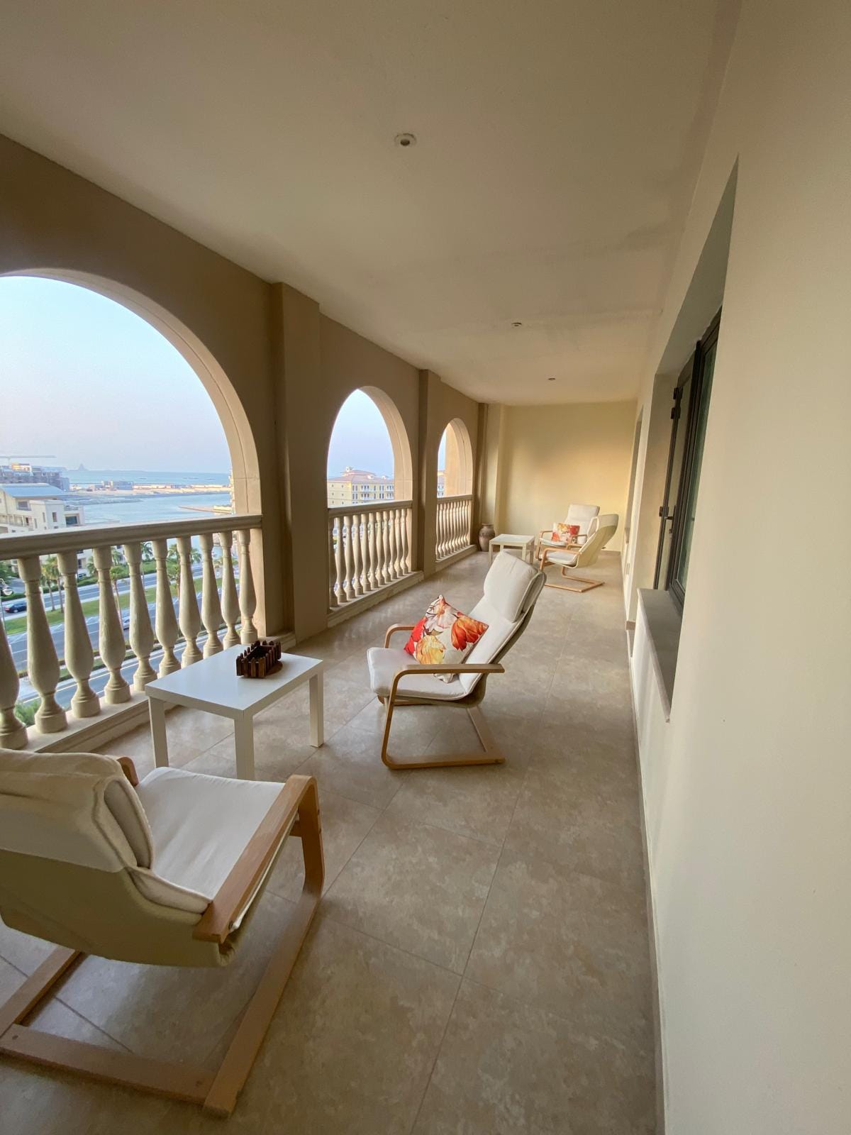 A spacious balcony is lined with arched openings, providing a scenic view. Comfortable seating arrangements are offered, featuring white chairs and small tables. Neutral walls and tiled flooring create a calm setting, inviting relaxation while enjoying the surroundings.