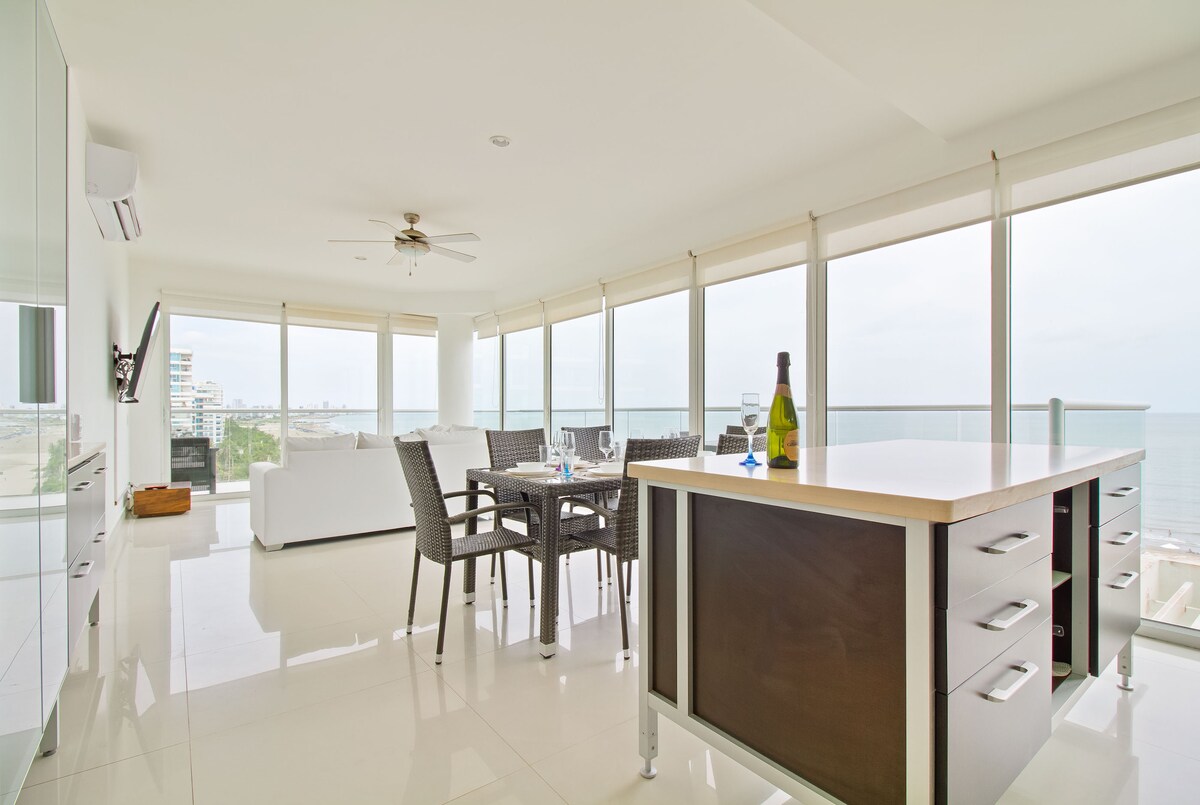 A bright and airy dining area features large windows offering stunning ocean views. A modern kitchen island is complemented by sleek cabinetry. Comfortable outdoor furniture is set for dining, while a ceiling fan enhances air circulation, creating a pleasant and welcoming environment.