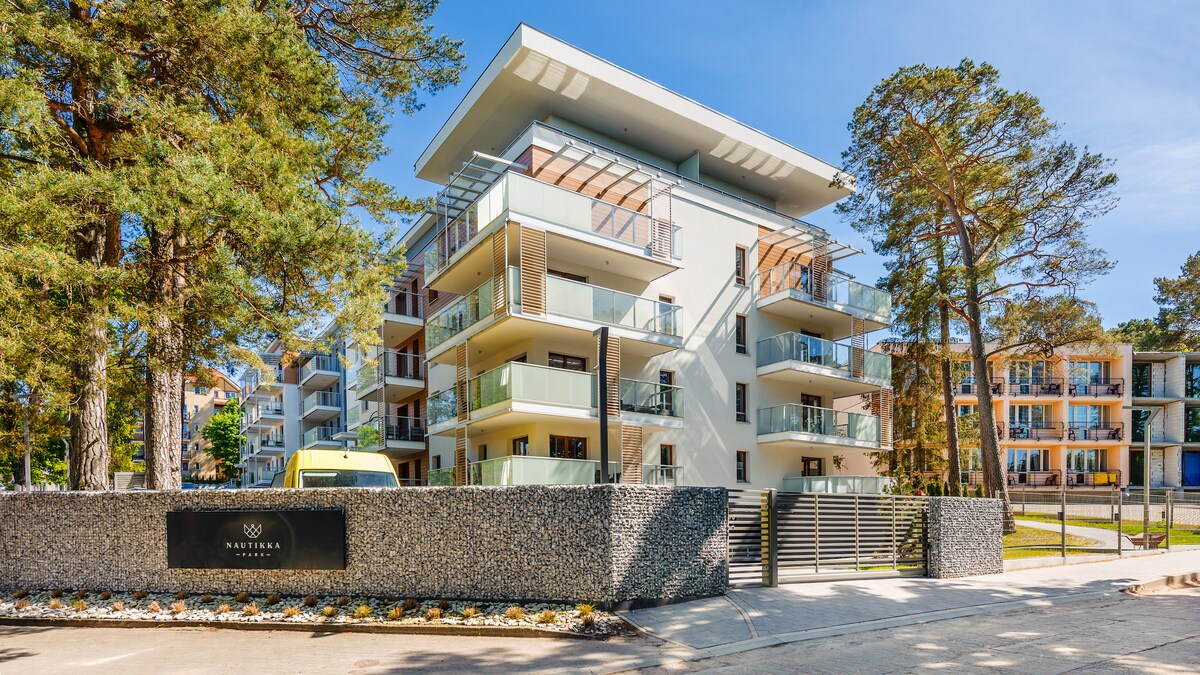 A modern multi-story building is surrounded by tall trees, featuring multiple balconies on each floor. The entrance is marked by a sleek stone wall displaying the property name. Clear blue skies and landscaped greenery enhance the serene outdoor setting.