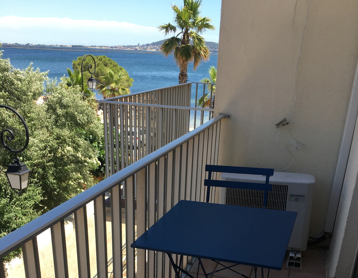 A small balcony is presented, featuring a blue table and chairs, with a view overlooking a body of water. Palm trees and greenery frame the scene, while distant buildings are visible along the shoreline under a clear blue sky.