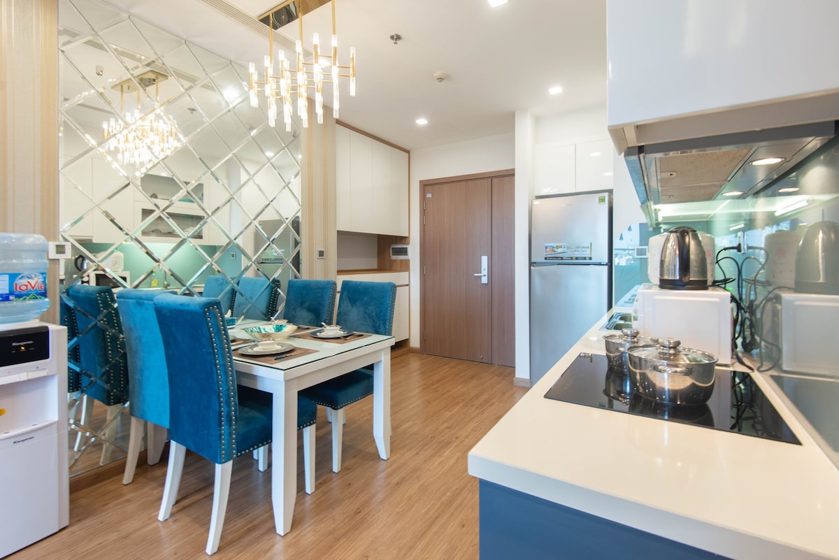 A modern kitchen and dining area are featured, highlighted by a glass mirror wall. A spacious table with six blue chairs is set for a meal. Stainless steel appliances, including a refrigerator and kettle, are visible, while a sleek countertop is equipped with cooking utensils.