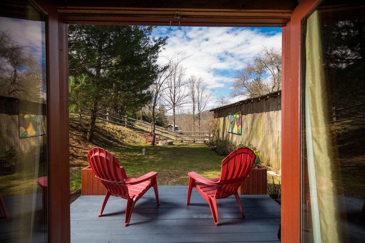 Two red Adirondack chairs are positioned on a porch, facing a grassy yard bordered by trees. In the background, a wooden fence complements the rustic setting, and colorful artwork is visible on the walls of nearby structures.