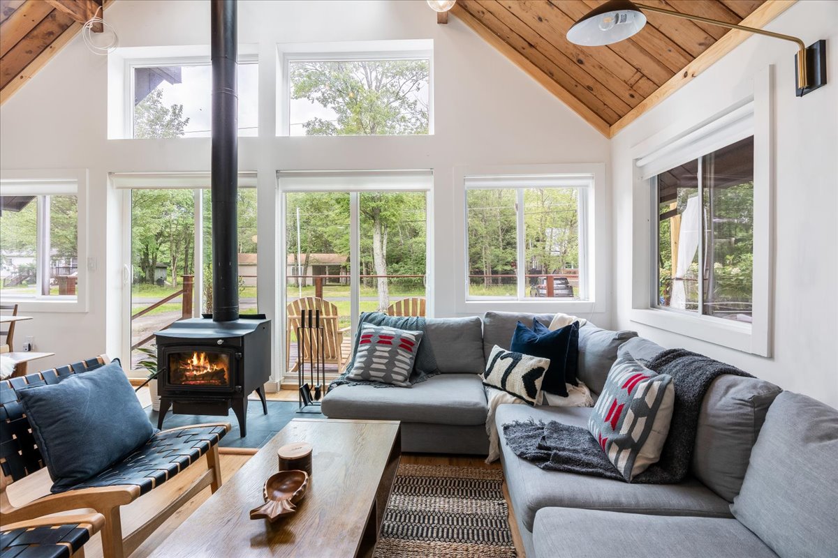 A cozy living area is highlighted by large windows that provide views of the wooded surroundings. A wood-burning stove stands centrally, complementing a spacious sectional sofa adorned with decorative cushions. Sunlight illuminates the room, enhancing the welcoming atmosphere.