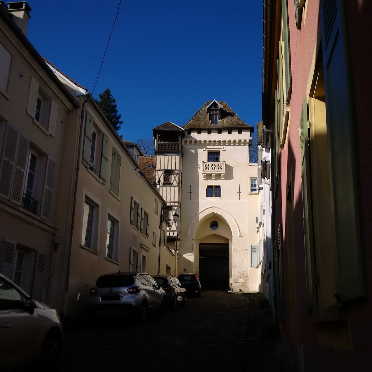 A cobblestone street lined with houses of varying colors leads towards an archway, showcasing an ancient structure with a distinctive tower above. The bright blue sky provides a clear backdrop, highlighting the architectural details and creating a sense of tranquility.