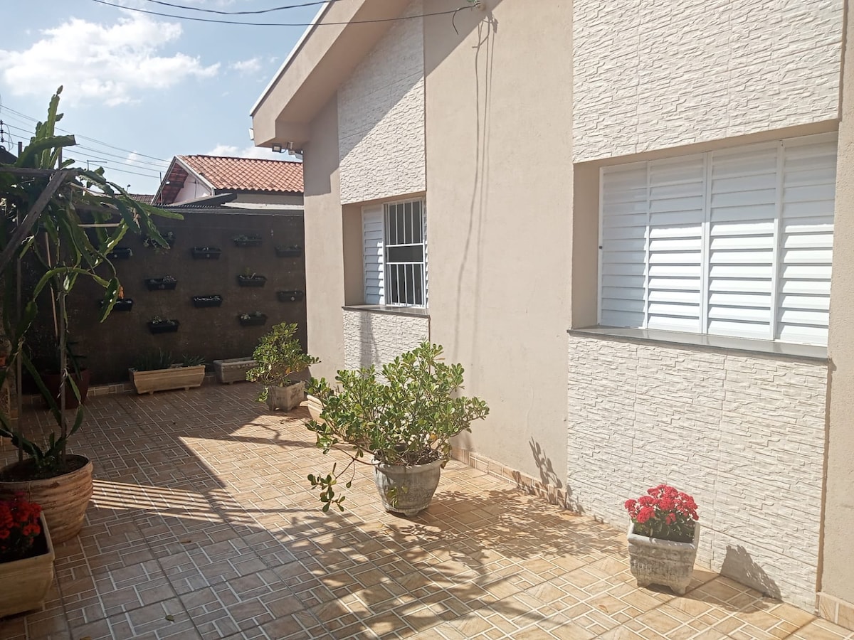 A well-maintained exterior features a paved patio area surrounded by several planters containing vibrant flowering plants. Two windows allow natural light to enter the space, with a textured wall adding visual interest. A vertical garden with additional plants is visible in the background.