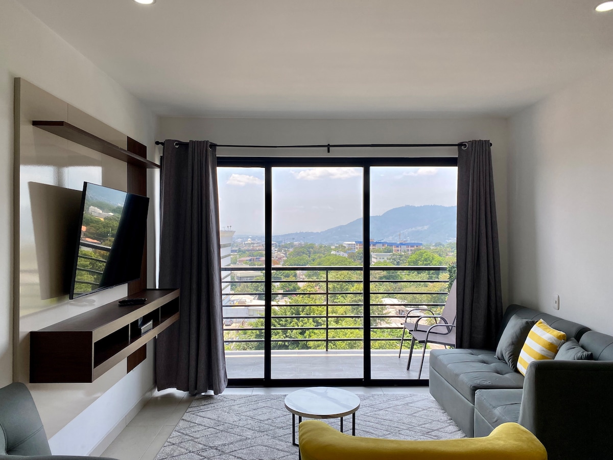 A modern living area is presented with a large window offering a scenic view of nearby mountains and the city. A comfortable sofa and a small coffee table create a space for relaxation, while a wall-mounted television adds convenience.