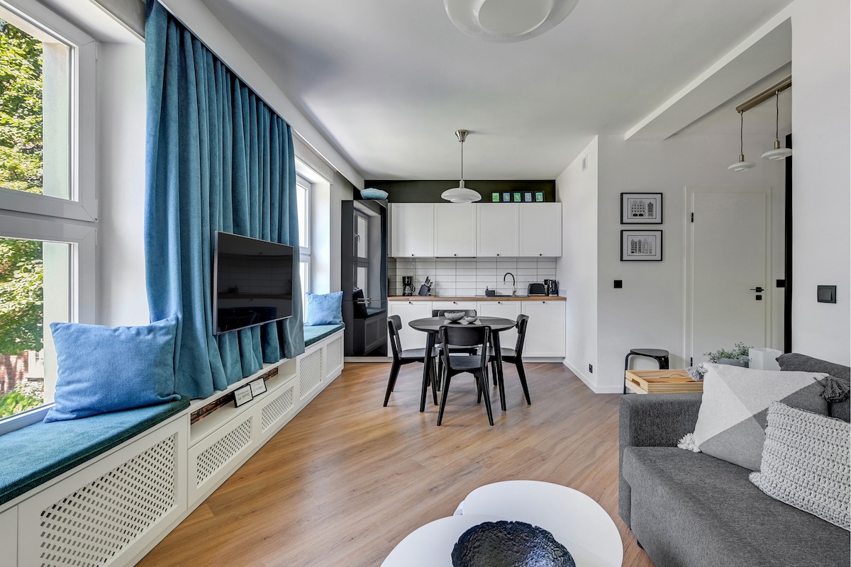 A modern living area is highlighted by large windows with blue curtains allowing natural light to fill the space. A dark sofa and stylish coffee table complement a dining area with a black table and chairs. A well-equipped kitchen is visible in the background.