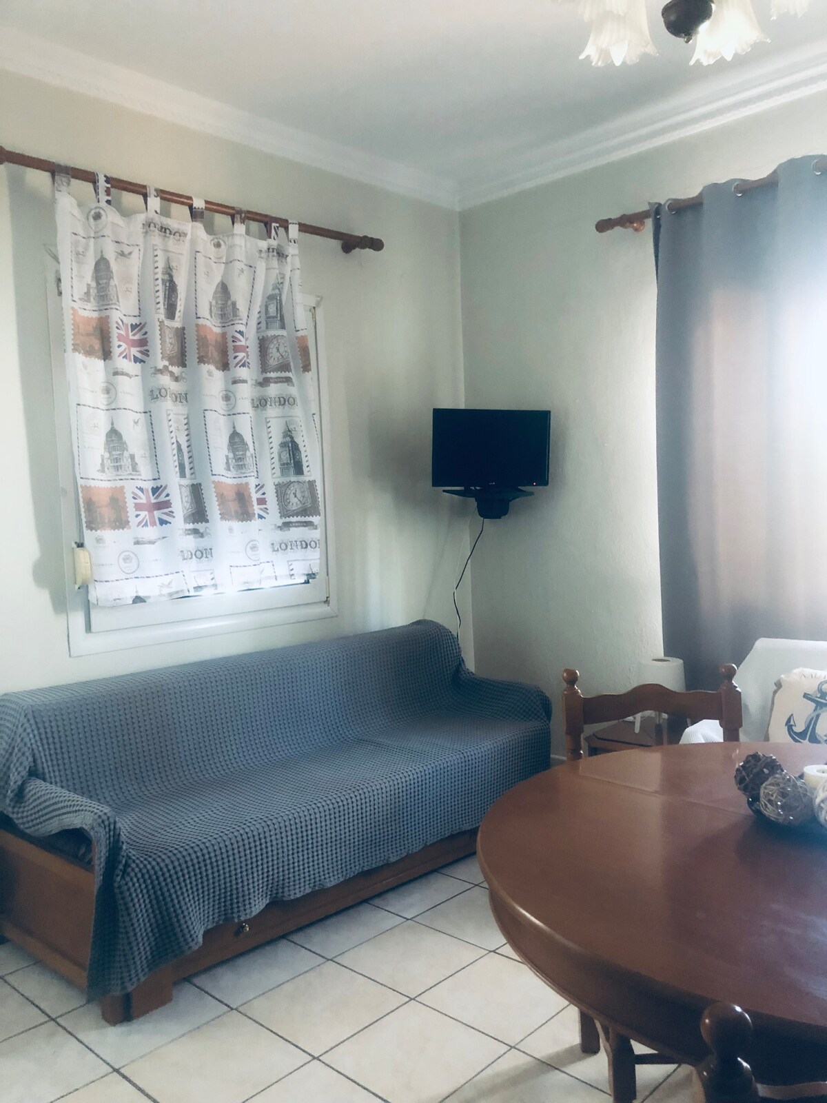 A cozy dining area features a round wooden table surrounded by four chairs. A comfortable sofa draped with a checkered blanket is positioned against the wall. Natural light enters through a window covered by a decorative curtain.