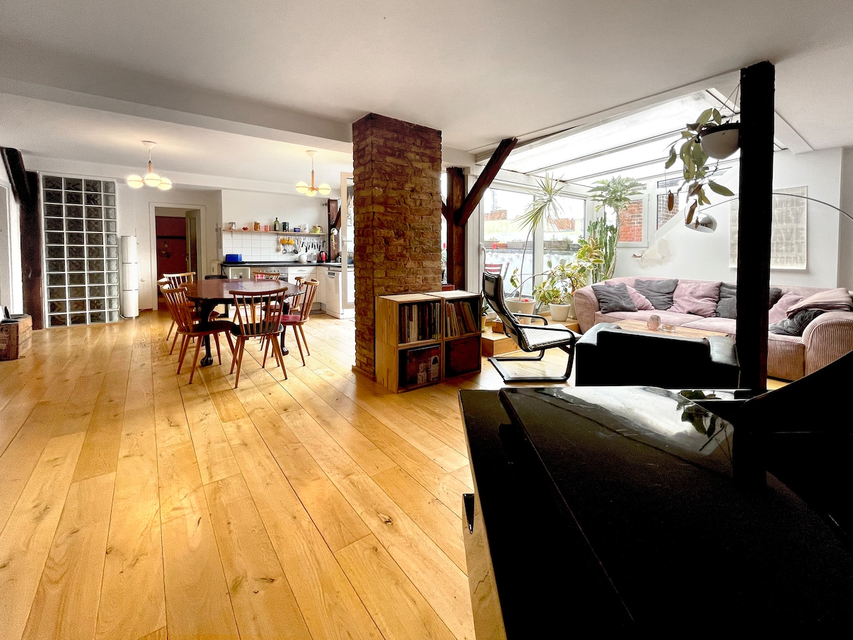 A spacious open-plan living area features warm wood flooring and exposed beams. A circular dining table with wooden chairs is situated near a brick pillar. Comfortable seating is arranged in a cozy corner, creating an inviting space for relaxation and socializing.