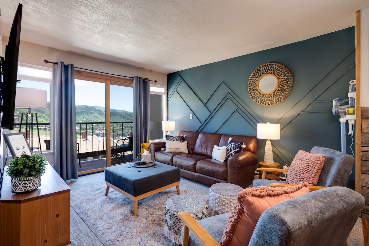 A cozy living area features a brown leather sofa and a blue accent wall with geometric patterns. Two lamps illuminate the space, complementing a coffee table and a decorative side table. Large sliding glass doors open to a balcony, offering scenic views of the surrounding hills.