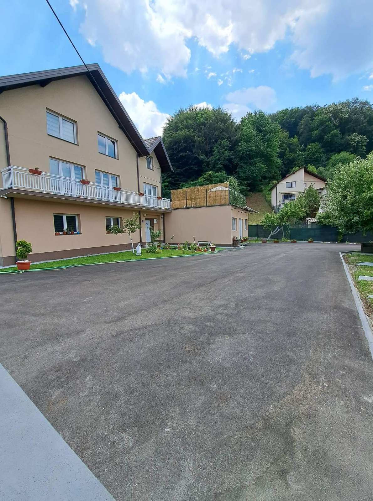 A spacious exterior view of the property is presented, featuring a well-maintained driveway leading to the entrance. Greenery surrounds the building, and a small garden area is visible, adding to the serene environment. The structure is set against a lush, wooded backdrop.