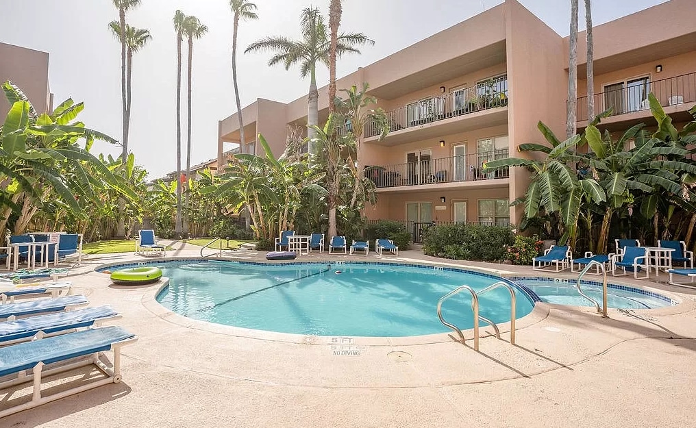 A heated pool is surrounded by lounge chairs and palm trees, providing a relaxing atmosphere. The pool area is bordered by well-maintained greenery and features a few steps leading to the water. The building exterior is visible in the background, with several balconies showcasing outdoor seating.
