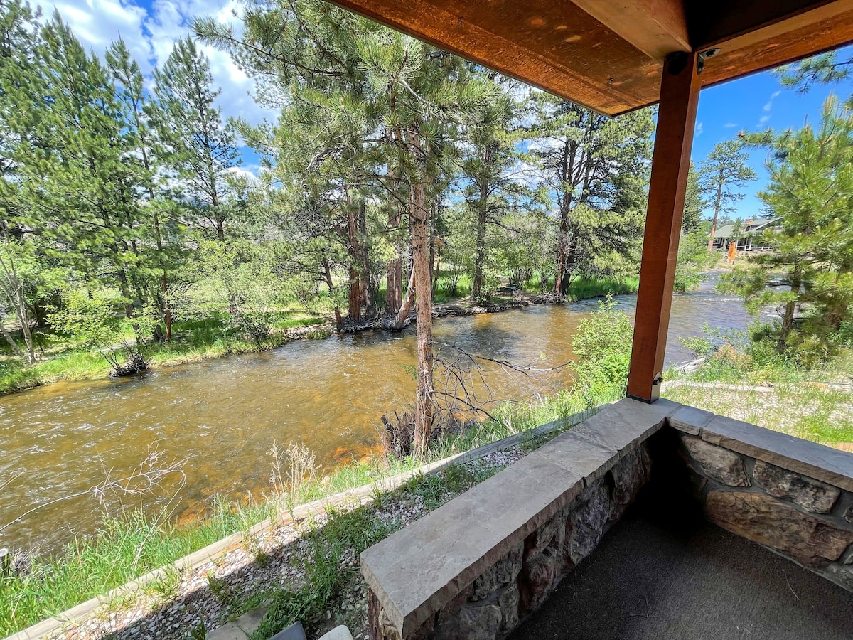 A shaded outdoor area features a stone seating ledge overlooking the Big Thompson River. Lush greenery is visible along the riverbanks, with trees framing the peaceful waterway under a bright sky.