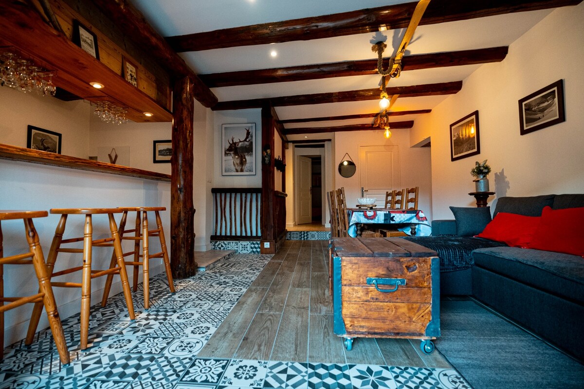 The open living area features rustic wooden beams and a patterned floor. A cozy seating arrangement includes a dark couch with a red accent pillow. A wooden bar with stools invites socializing, while decorative wall art and lighting enhance the welcoming ambiance.