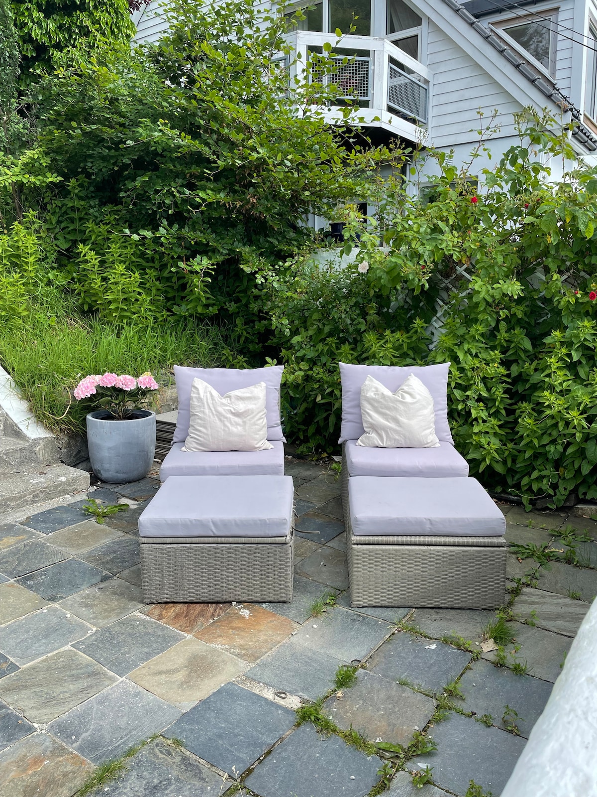 Two outdoor loungers are positioned against a backdrop of lush greenery. Each lounger features soft pillows for comfort. A potted plant with pink flowers adds a touch of color beside the loungers, set on a natural stone patio that creates a serene atmosphere.