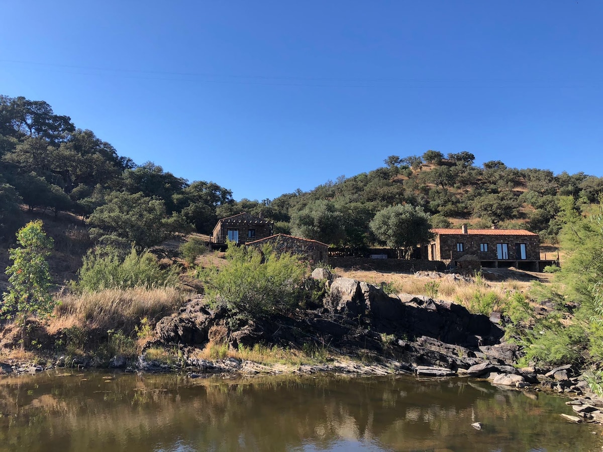 Two charming houses are nestled on a hillside, surrounded by lush green vegetation. The structures display earthy tones and are complemented by a clear blue sky. A calm river flows at the foreground, reflecting the tranquil scenery.