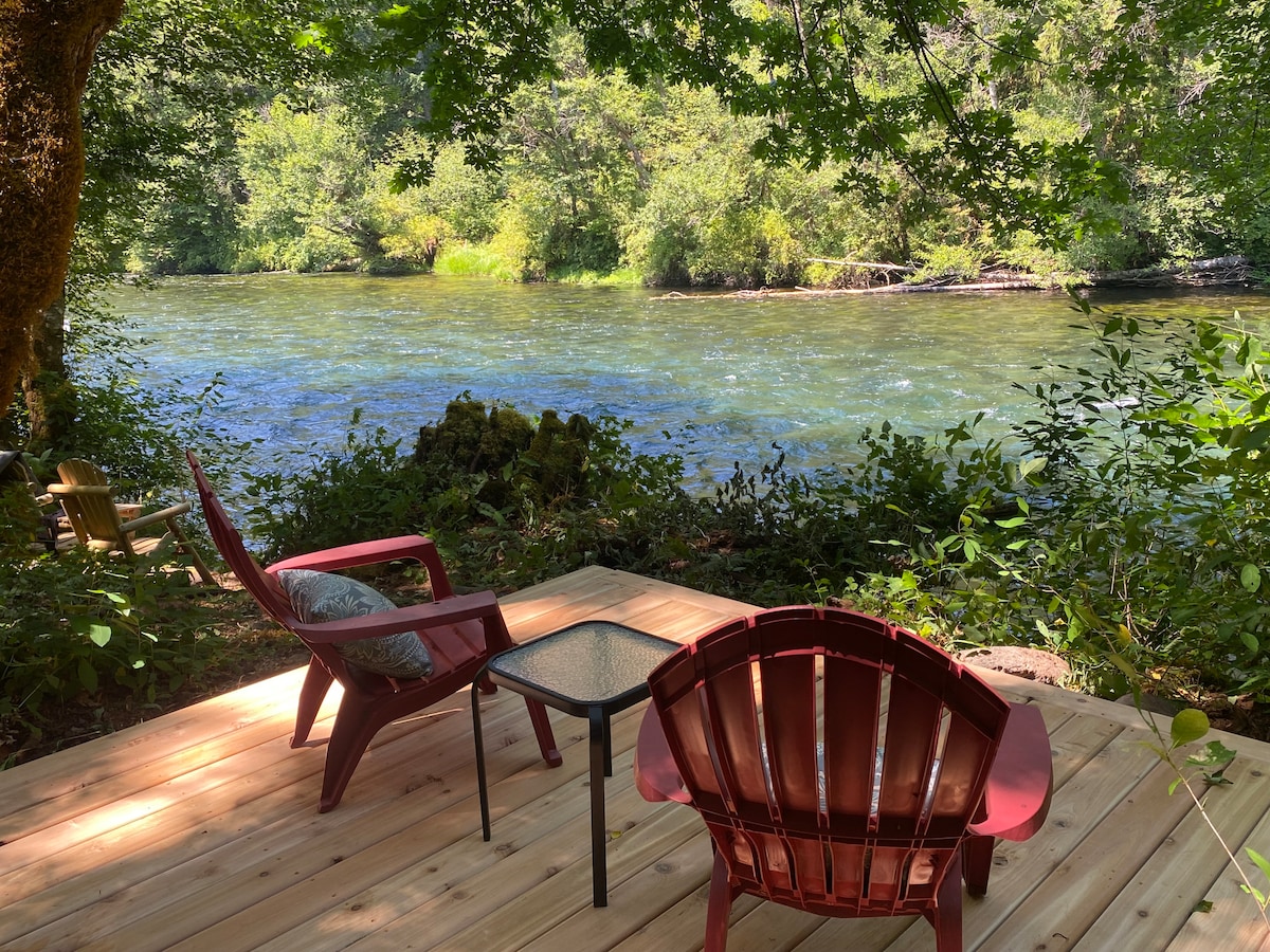 A wooden deck features two red Adirondack chairs and a small table, set along the bank of the McKenzie River. Lush greenery surrounds the area, while clear waters flow gently past, creating a serene and inviting outdoor space.