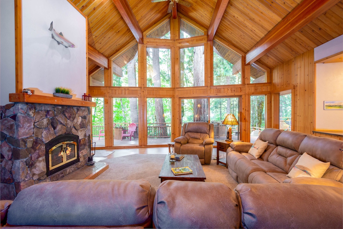 The living room features high wood-paneled ceilings, with large floor-to-ceiling windows that provide views of the surrounding forest. A stone fireplace is centered between two comfortable reclining couches, while a wooden coffee table is positioned in front, creating a cozy seating arrangement.