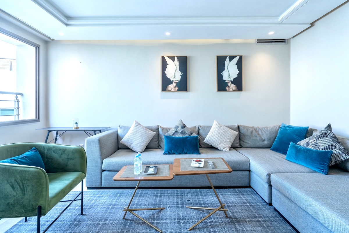 The spacious living room features a large, comfortable sectional sofa adorned with various cushions in shades of grey and blue. Two framed artworks are displayed on the wall above a stylish gold-accented coffee table, enhancing the room's modern aesthetic.