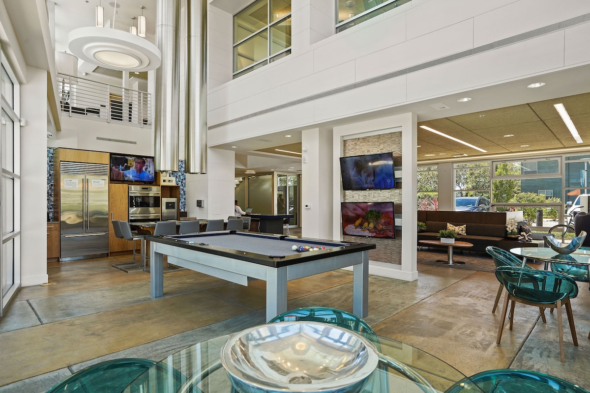 A welcoming communal space features high ceilings and large windows that allow natural light to fill the area. A billiards table is positioned centrally, surrounded by comfortable seating. Several flat-screen TVs are mounted on the walls, enhancing the recreational atmosphere.